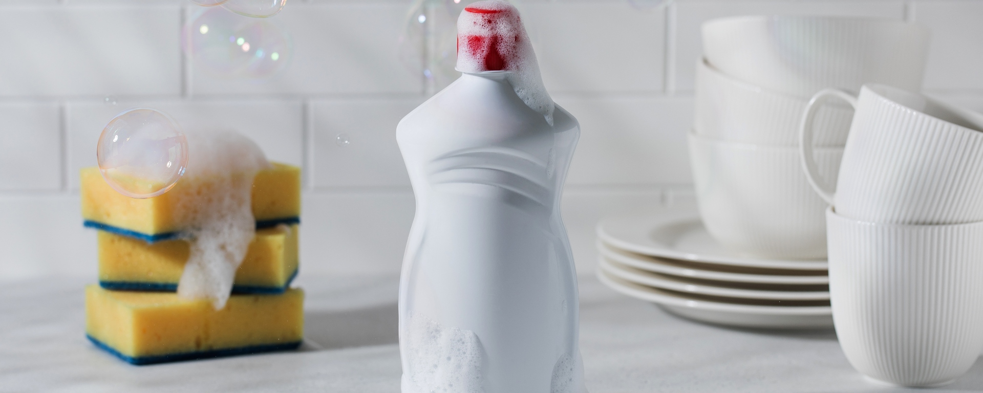 Bottle of dishwashing liquid in between sponges and dishes, with bubbles in the air